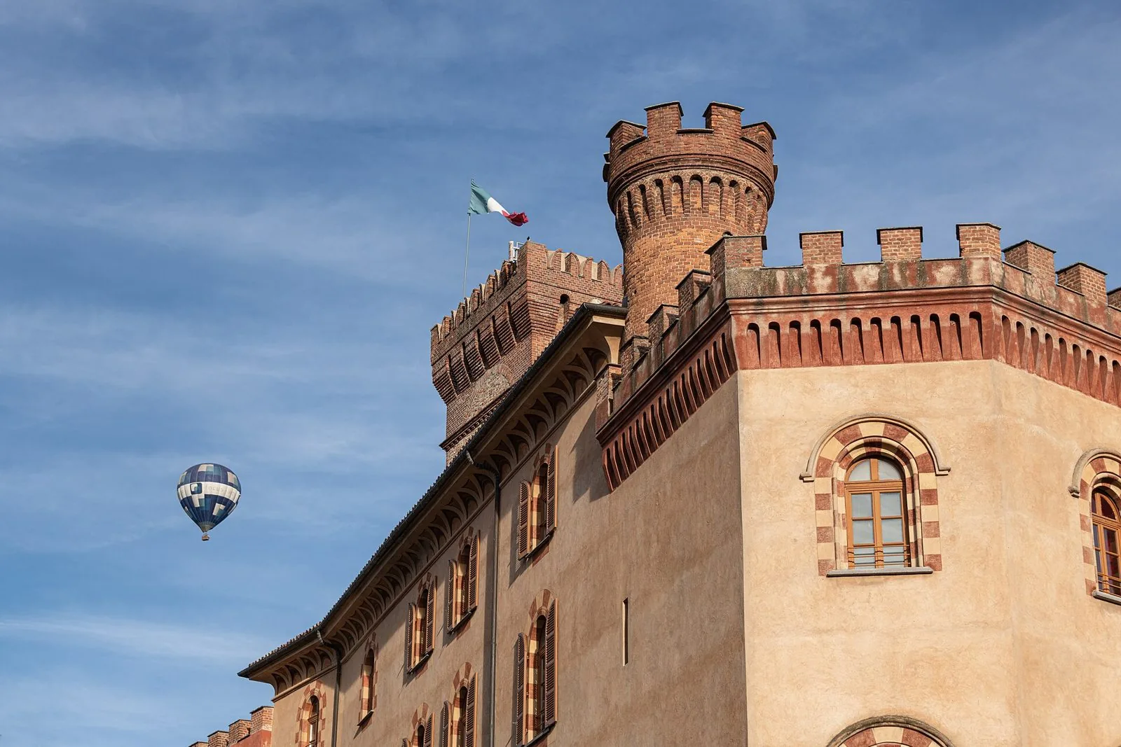 Le torri merlate in mattoni e la facciata del castello Falletti di Barolo contro il cielo azzurro, con una mongolfiera in volo sullo sfondo